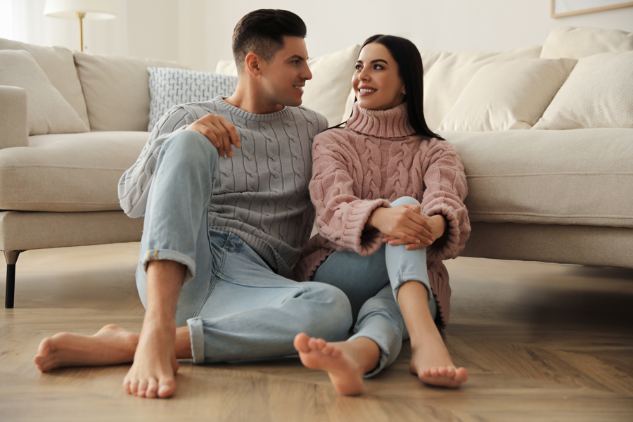 A couple sits on the floor, relaxed and engaged, in front of a cozy couch in a warm living room setting